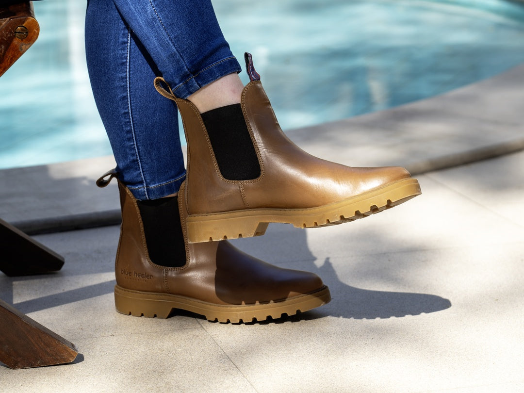 A person wearing tan leather Chelsea-style boots with black elastic side panels and tan lug soles, shown outdoors by a pool with blue jeans visible.