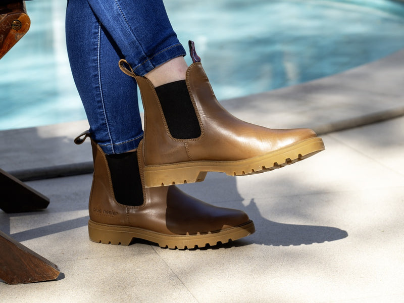A person wearing tan leather Chelsea-style boots with black elastic side panels and tan lug soles, shown outdoors by a pool with blue jeans visible.