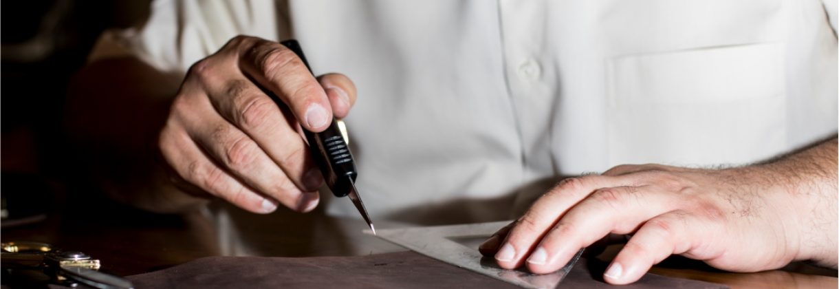 Close-up of artisan hand-cutting brown leather sheet using precision knife and ruler for handmade boot crafting.